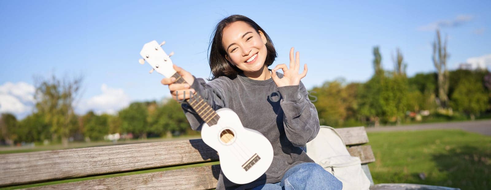 Cute smiling girl shows ok sign and her new ukulele, sits on bench in park, recommends musical instrument.