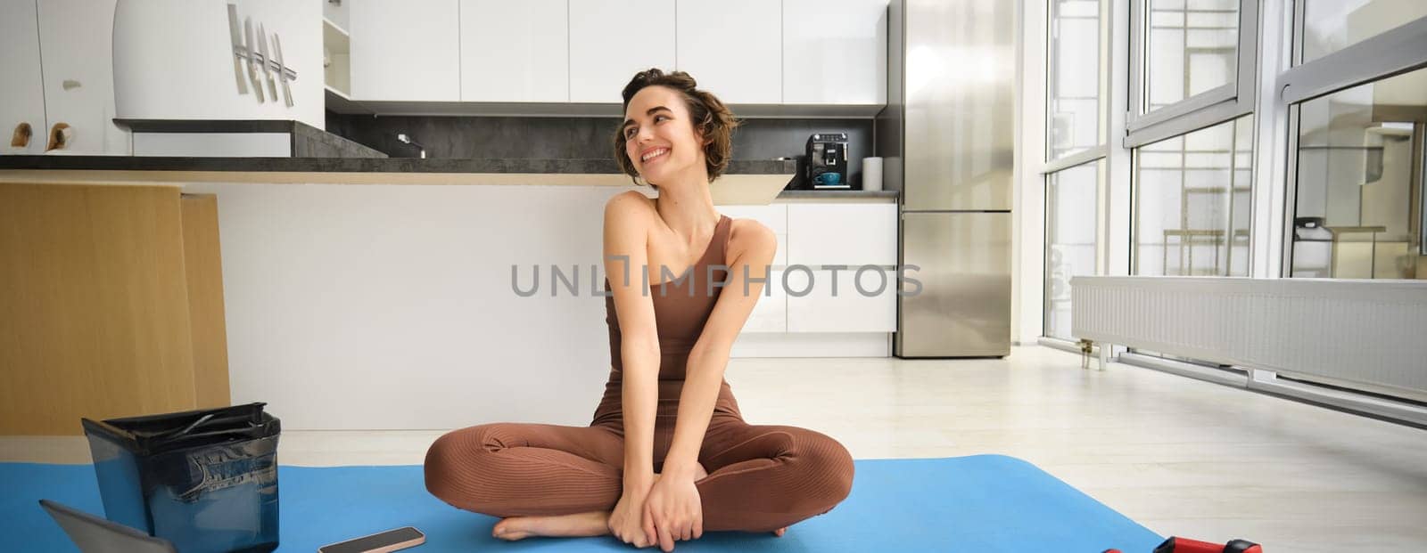 Sport and wellbeing. Young brunette fitness girl, wearing activewear, sitting on yoga mat, meditating, practice relaxation and mindfulness at home, workout in kitchen. Copy space