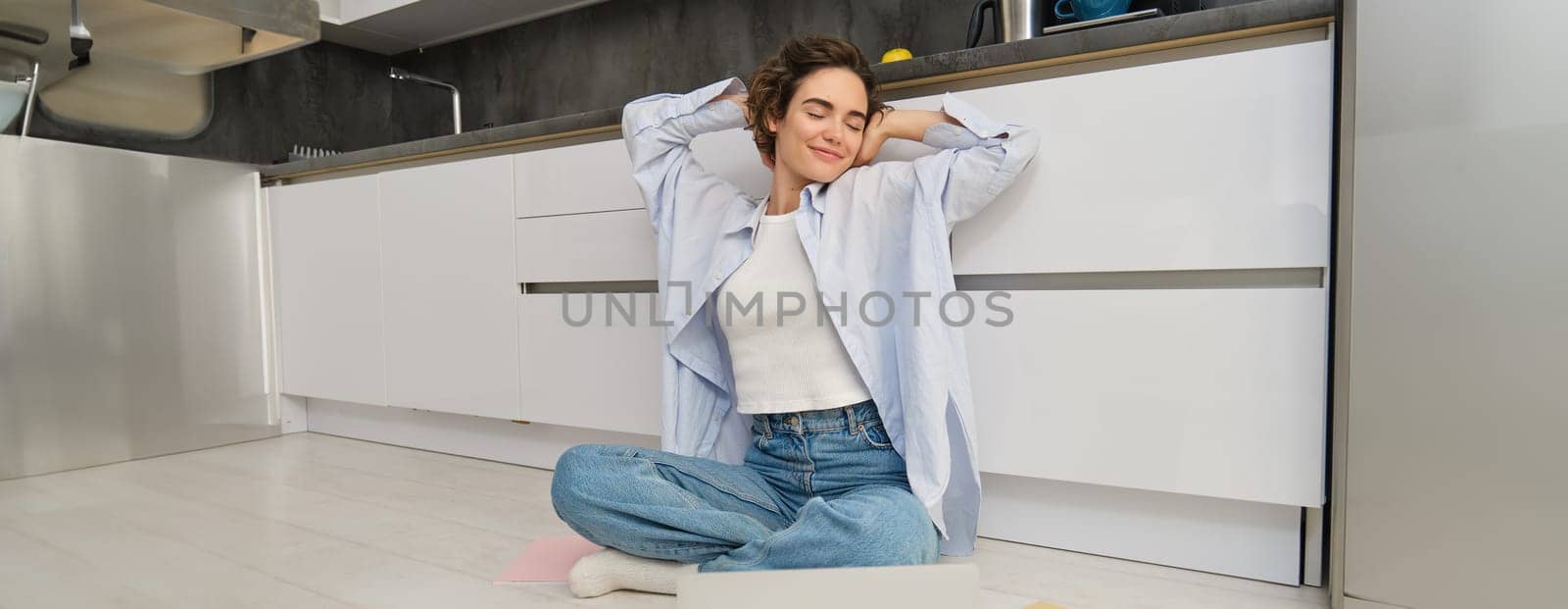 Happy young woman stretches her arms, sits on floor with laptop, finishes her remote work, completes homework on computer. Technology and people concept