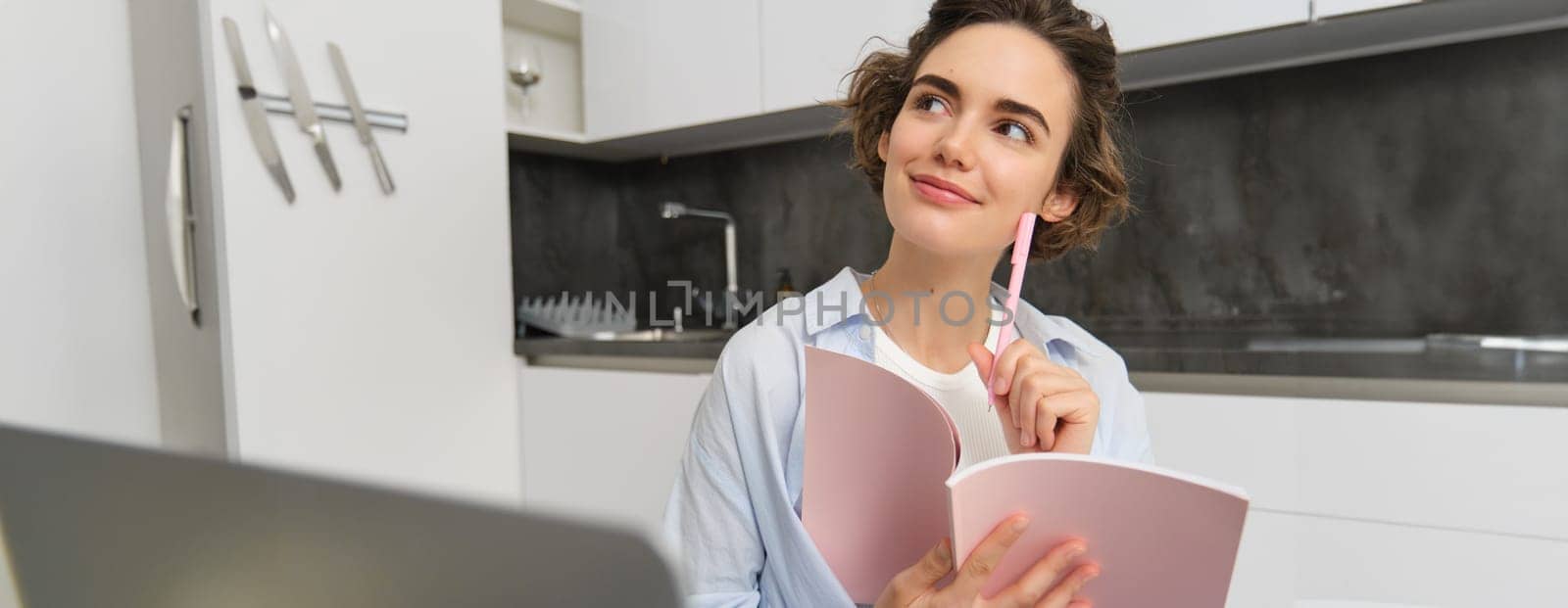 Portrait of young businesswoman, works from home, looks thoughtful while writes in planner, takes notes during online classes, noting information from webinar.