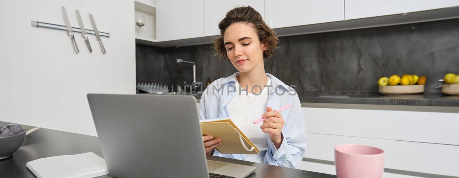 Working woman makes notes, writes down information on notebook, sitting with laptop at home by Benzoix