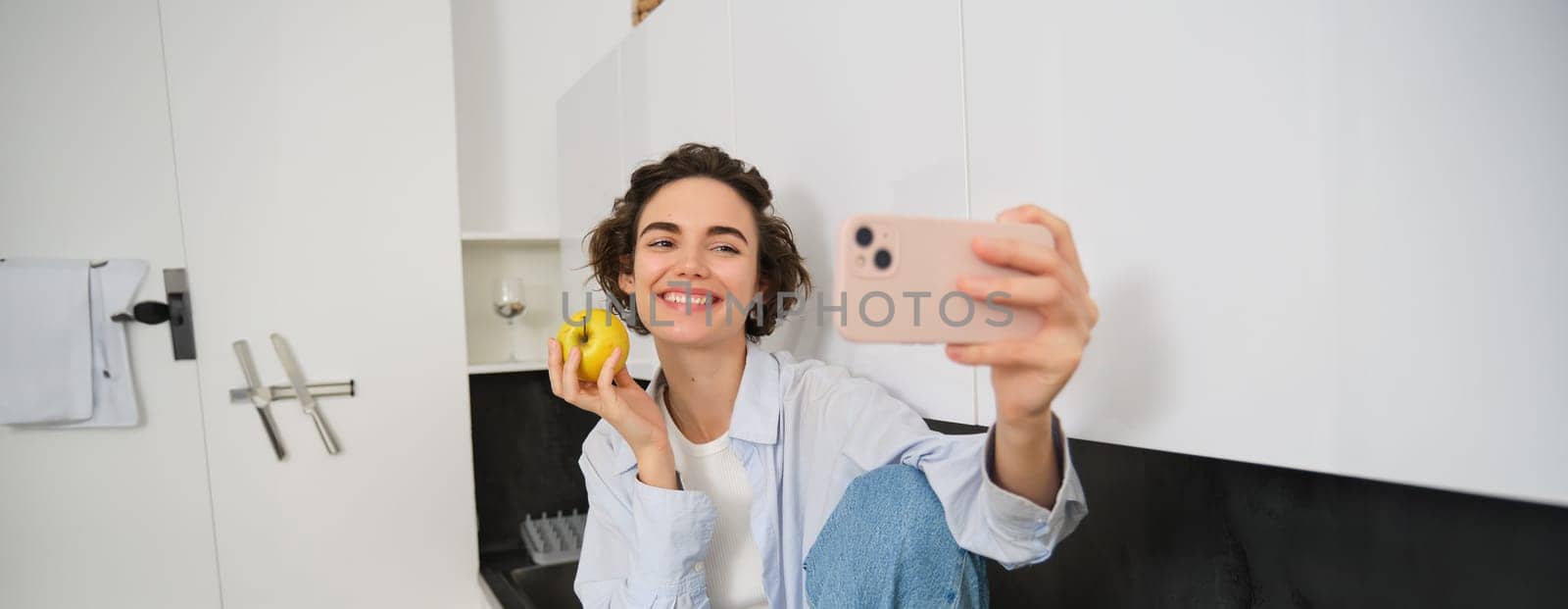 Beautiful girl blogger, takes selfie as she eats apple, shows how to lead healthy lifestyle to followers online, holds smartphone for photo.