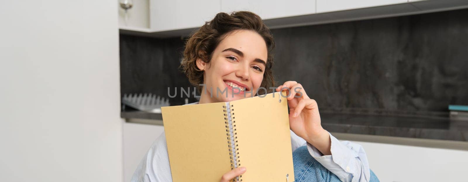 Image of brunette girl at home, holding notebook, reading through her notes, preparing for exam, doing homework in kitchen. Lifestyle and people concept