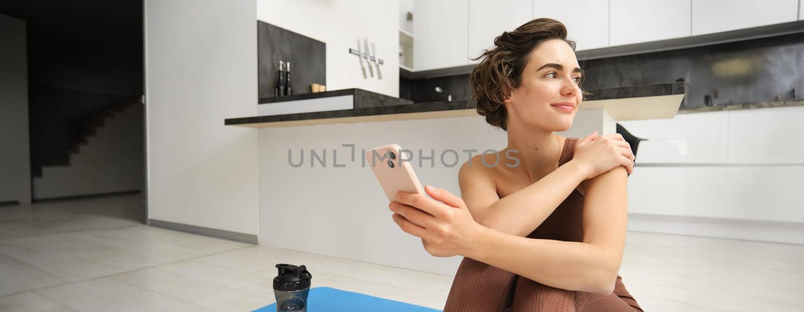 Portrait of sporty girl with mobile phone, sitting on yoga mat, using smartphone during workout at home, watching fitness turorial on telephone. Sport and wellbeing concept