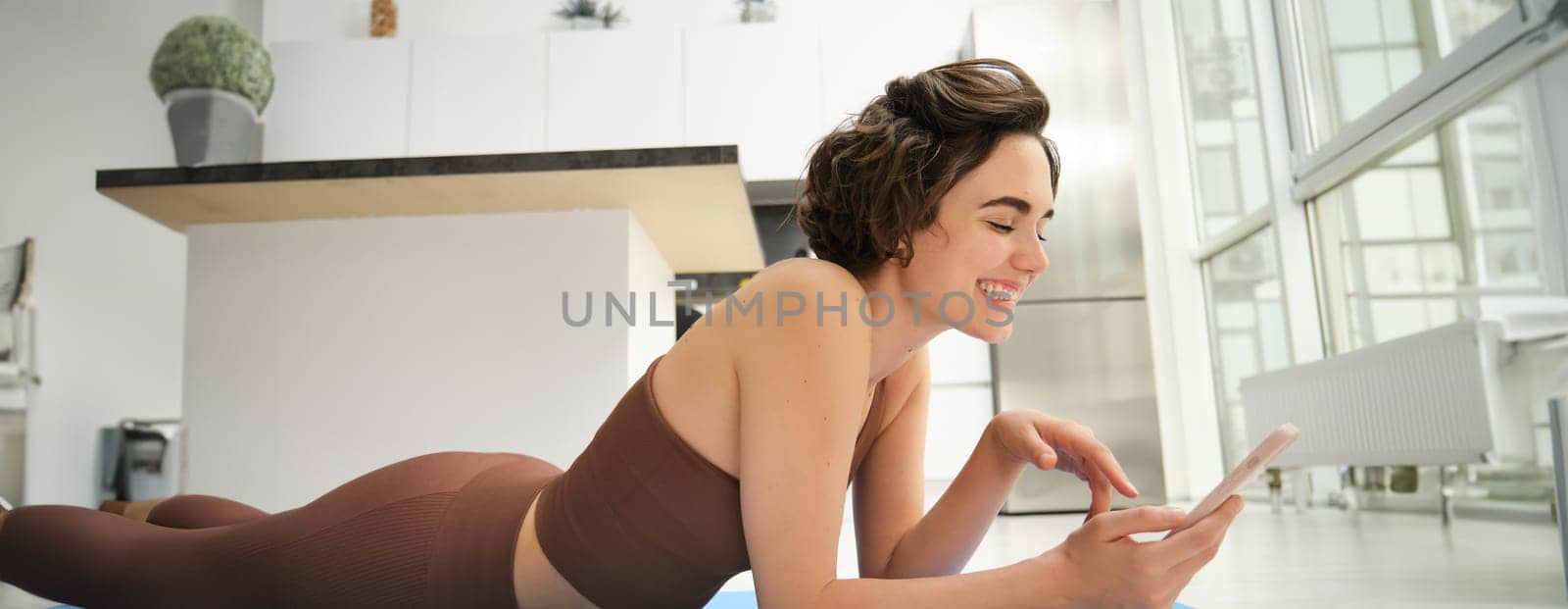 Fitness and technology. Smiling brunette woman in activewear, workout at home, looking at smartphone with happy face, watching training instructor on mobile phone app.