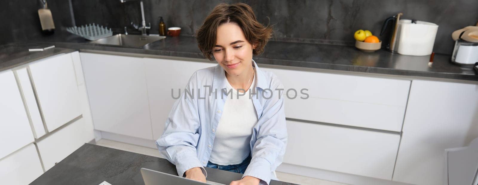 Young woman works from home, uses laptop in her kitchen. Student doing homework. Girl studies, watches online webinar, does distance learning. Courses and technology concept