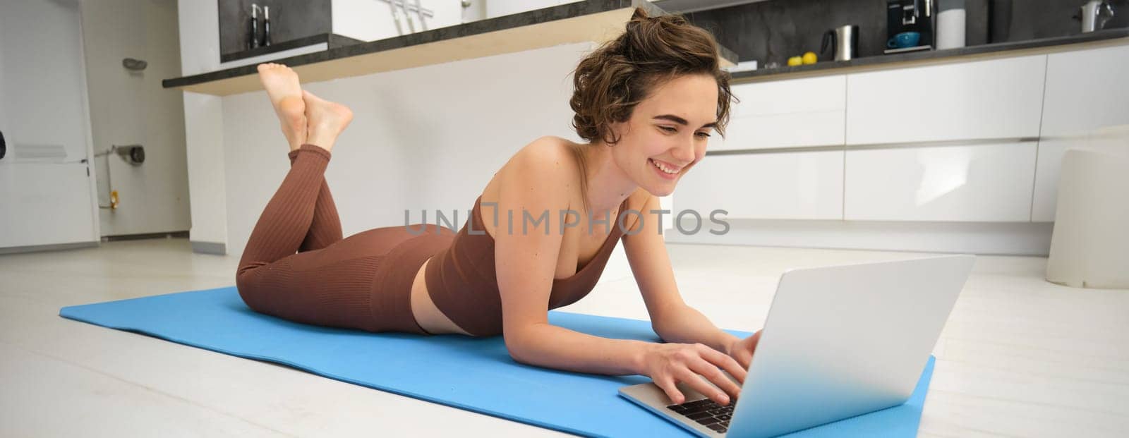 Portrait of woman athlete, girl workout at home, watches fitness videos on laptop, lying on rubber mat and doing indoor yoga session. Sport and wellbeing concept