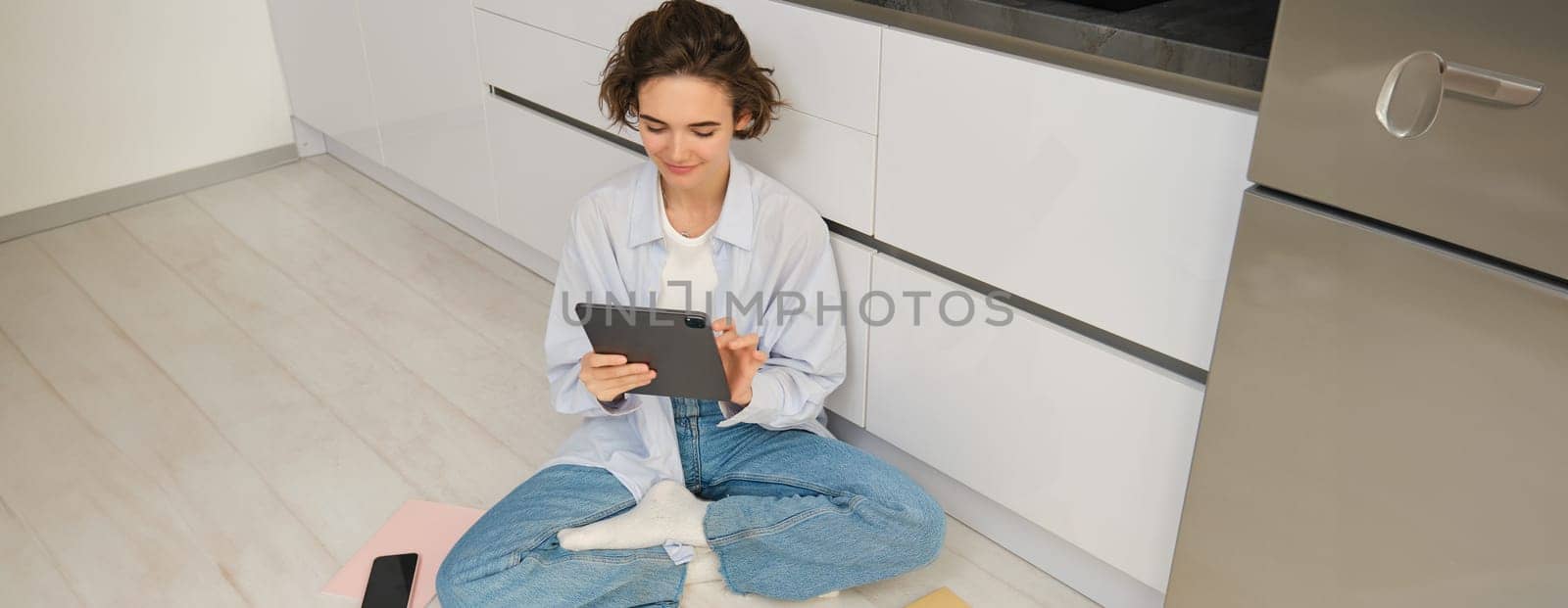 Young woman, freelancer works from home, looks at her digital tablet, reads through documents online, smiling and sitting on floor in kitchen.