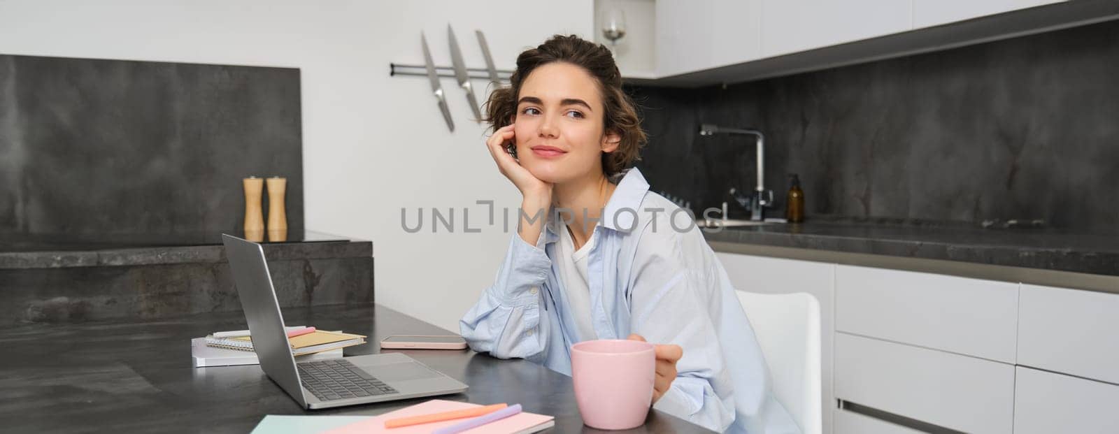 Beautiful brunette woman, looking outside window, working from home, drinking coffee in kitchen and suign laptop.