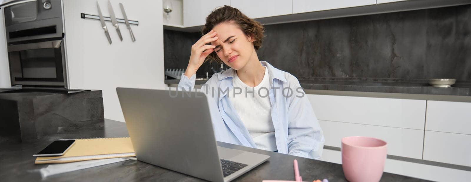 Woman has headache while works from home. Girl has painful migraine, sits near laptop, grimaces from discomfort.