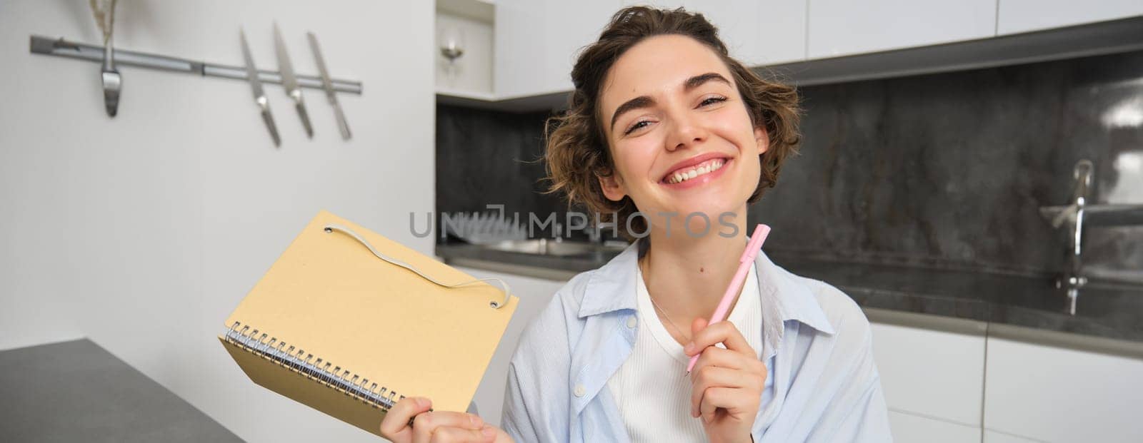 Portrait of smiling brunette girl, holds her planner, writes in notebook and looks happy, laughs, does homework, makes notes, sits at home in kitchen.