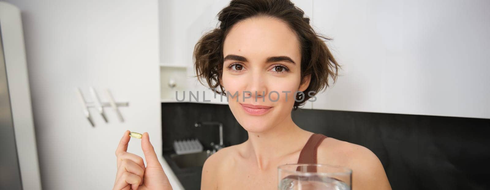 Portrait of sportswoman drinking water, taking vitamins, dietry supplements for healthy skin, having omega-3 pill, standing in her kitchen in workout clothing.