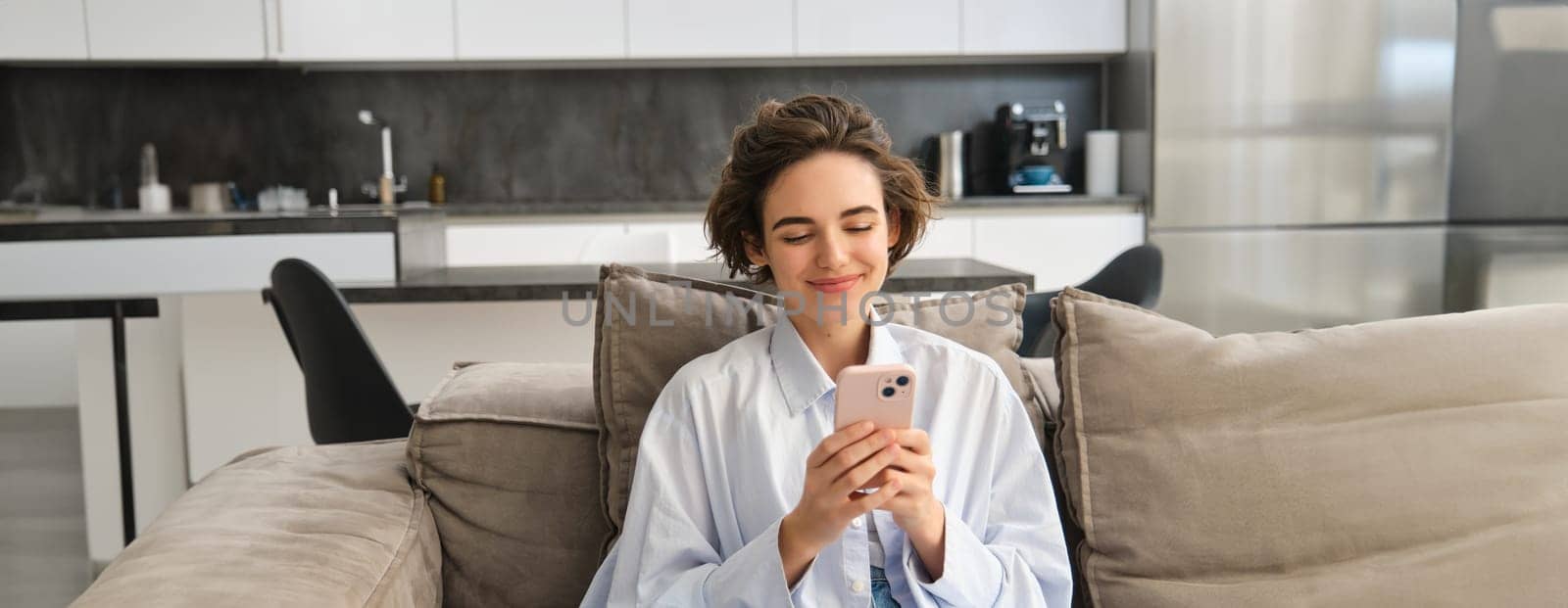 Image of happy woman sits on couch with smartphone, looks at mobile phone screen with pleased smile, chats in app, watches videos online.