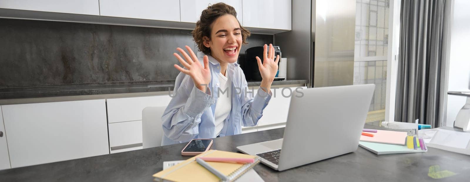 Beautiful girl waves hands at laptop camera, says hello to coworkers, connects to work meeting online, studies on remote from her kitchen by Benzoix