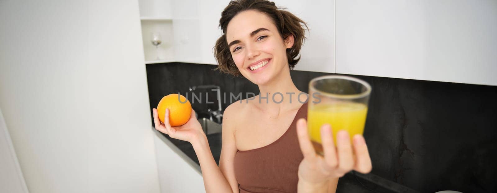 Healthy body and wellbeing. Smiling fitness girl giving you glass of fresh juice, holding an orange and looking happy. VItamin drink after workout at home.