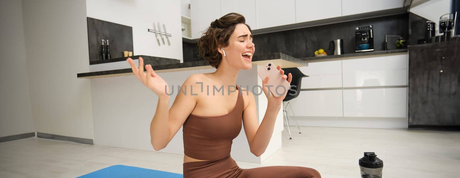 Carefree fitness girl, workout instructor sits at home with water bottle, yoga mat, wears tracksuit, sings into her smartphone, enjoys music in wireless headphones, listens song during pilates yoga by Benzoix