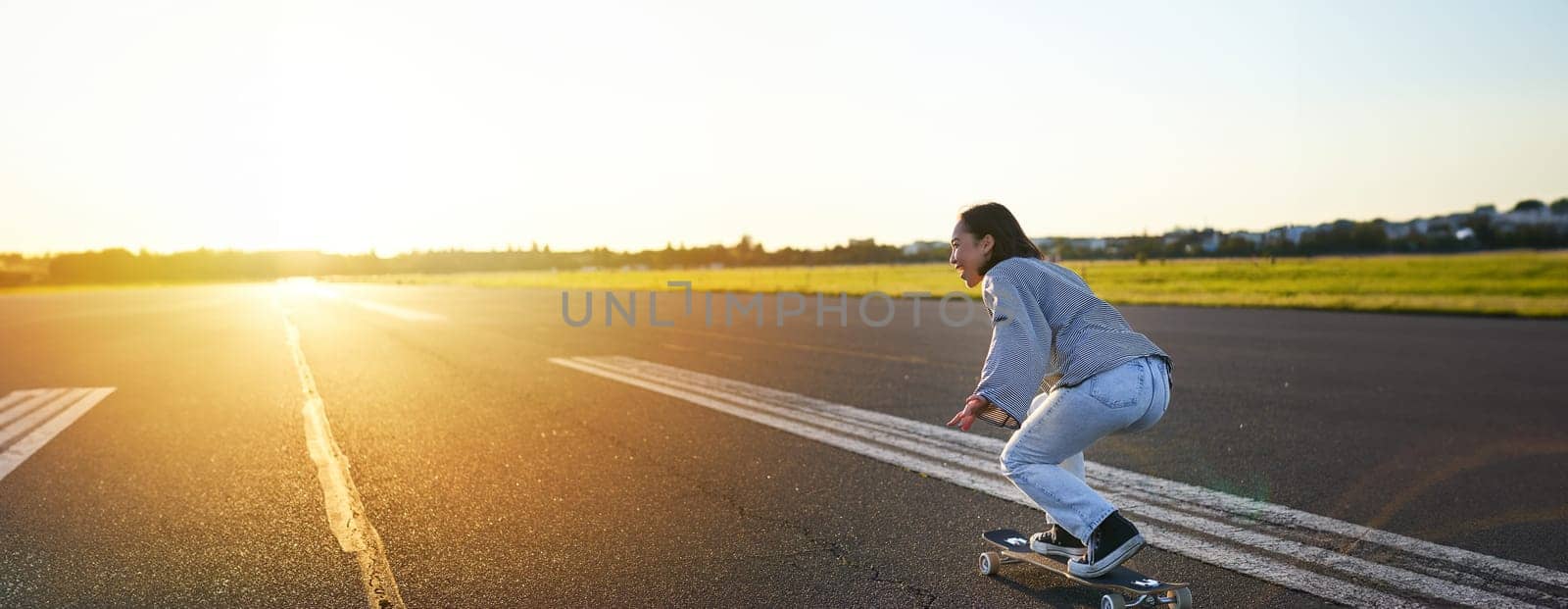 Side view of beautiful asian girl on skateboard, riding her cruiser towards the sun on an empty road. Happy young skater enjoying sunny day on her skate.