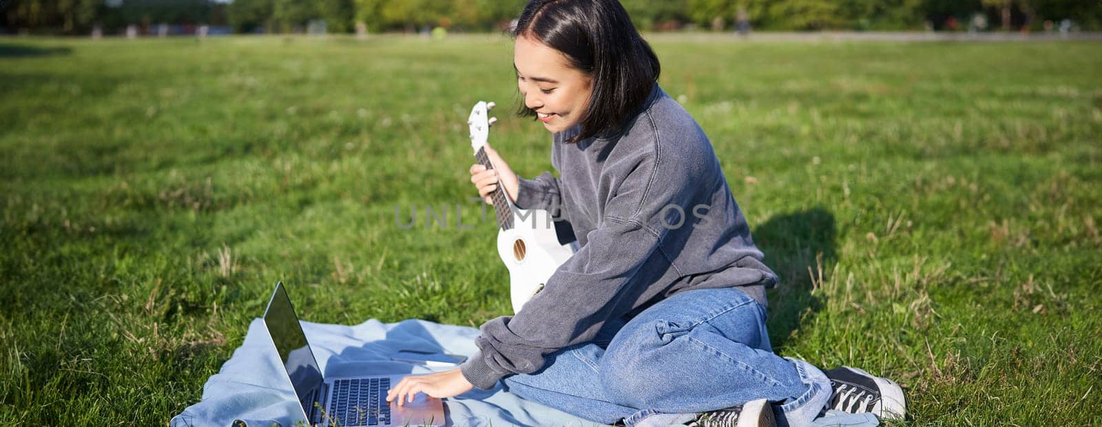 Smiling asian girl learns how to play ukulele via laptop, online video tutorials, sitting on grass in park with musical instrument.