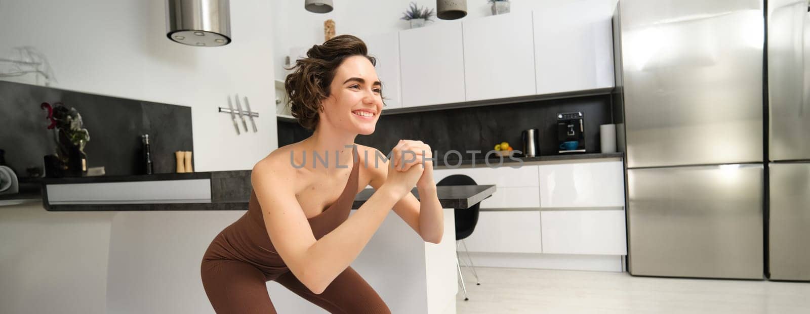 Image of young woman doing squats at home, workout on rubber mat in bright room indoors, wearing activewear for training. Copy space