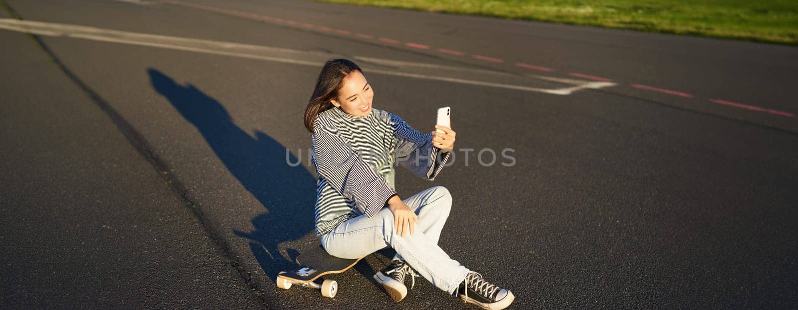 Beautiful korean girl takes selfie on smartphone, takes photo with her skateboard, enoys sunny day outdoors.