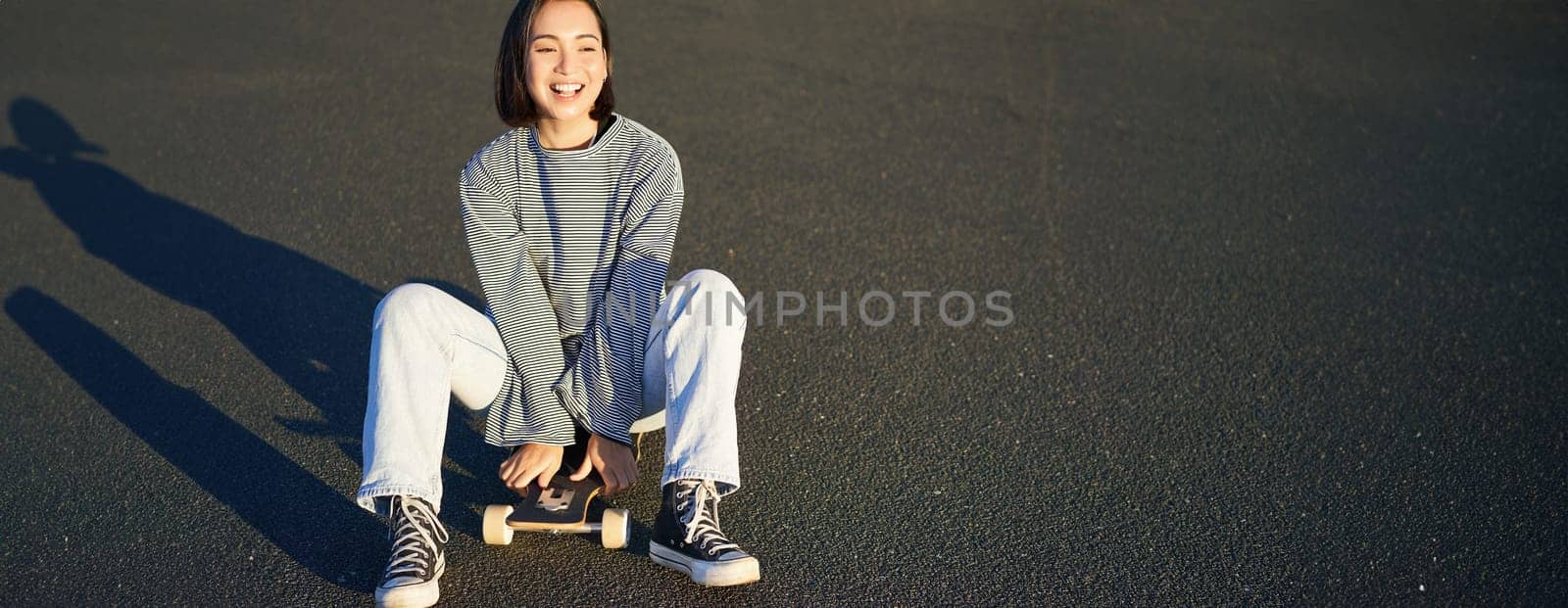 Freedom and happiness. Cute smiling asian girl, sits on skateboard on sunny spring day. Happy laughing skater enjoying cruising on longboard.