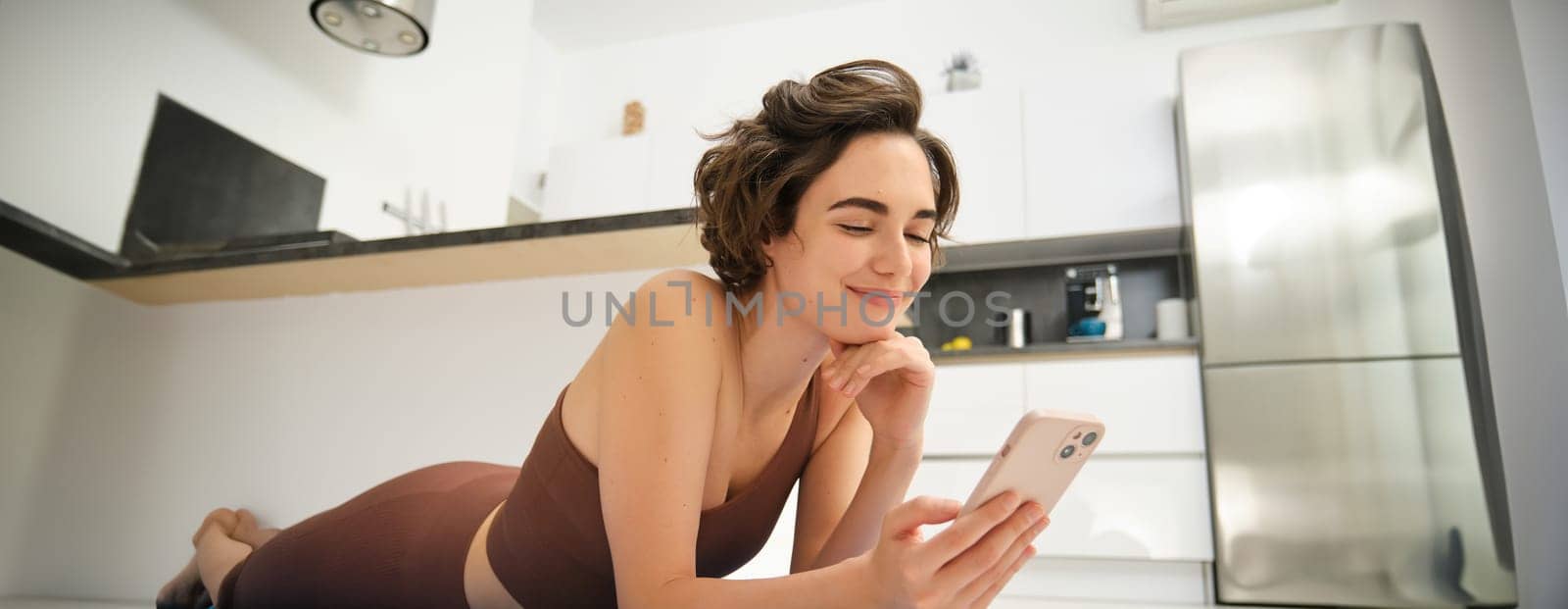 Image of sporty girl resting on yoga rubber mat, workout at home, using her smartphone, looking for online fitness tutorials, mobile app for training by Benzoix