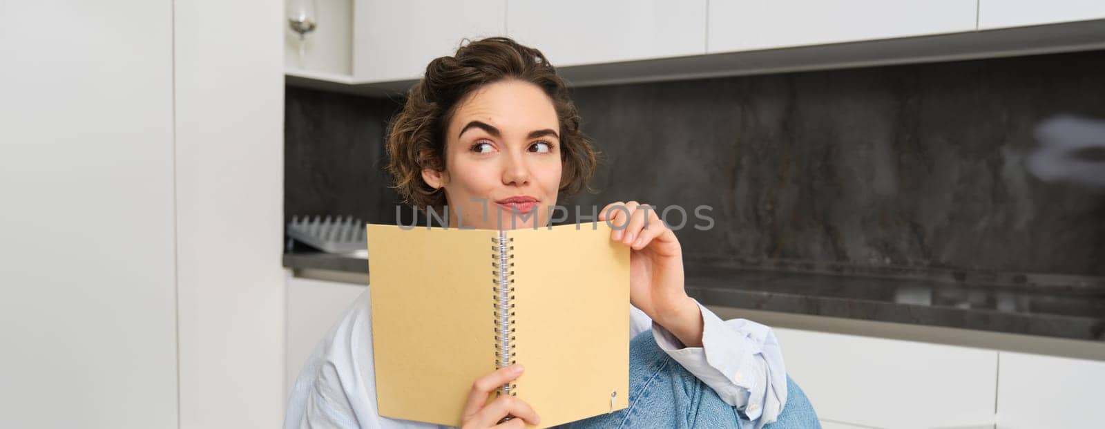 Portrait of beautiful young woman, looking intrigued, reading notes and smiling with thoughtful face, sitting at home in kitchen. Lifestyle and people concept