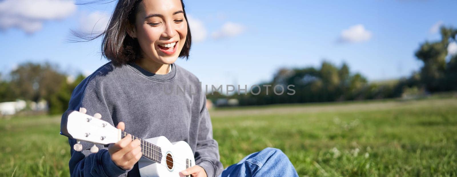 Happy people and hobbies. Smiling asian girl playing ukulele guitar and singing, sitting in park outdoors on blanket.