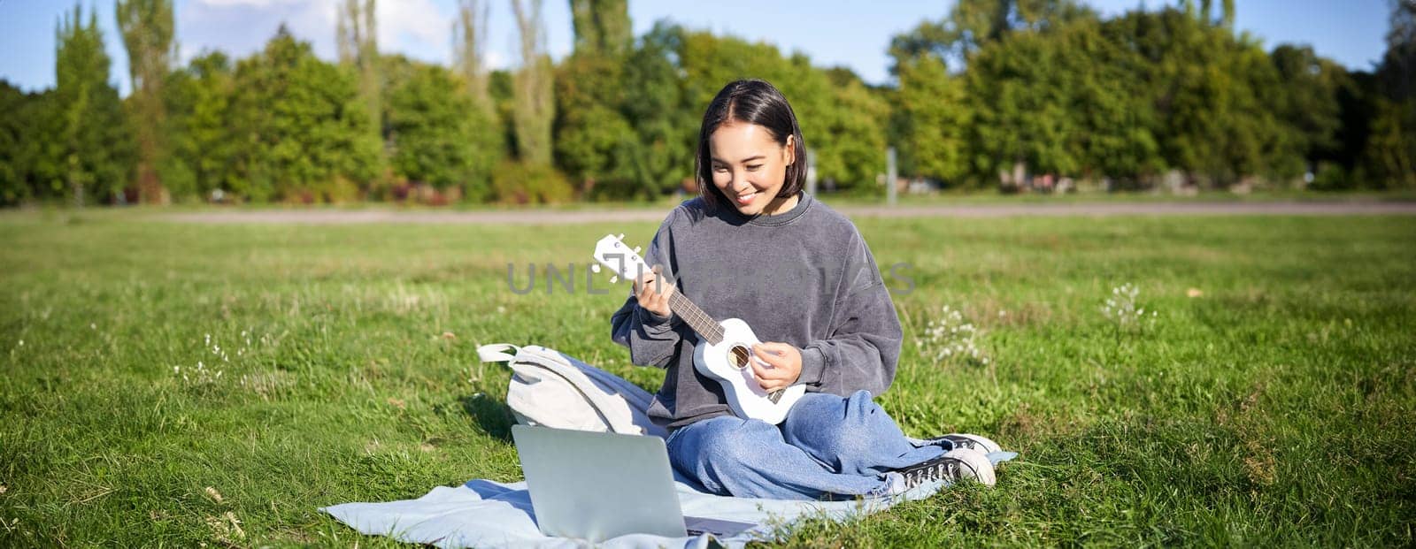 Happy asian girl plays ukulele outdoors, teaches music online with her laptop, sitting in park, playing instrument.