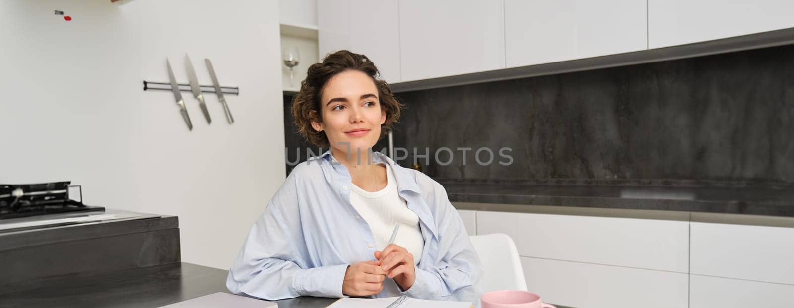 Portrait of young woman at home, working, writing in notebook, taking notes or doing homework by Benzoix