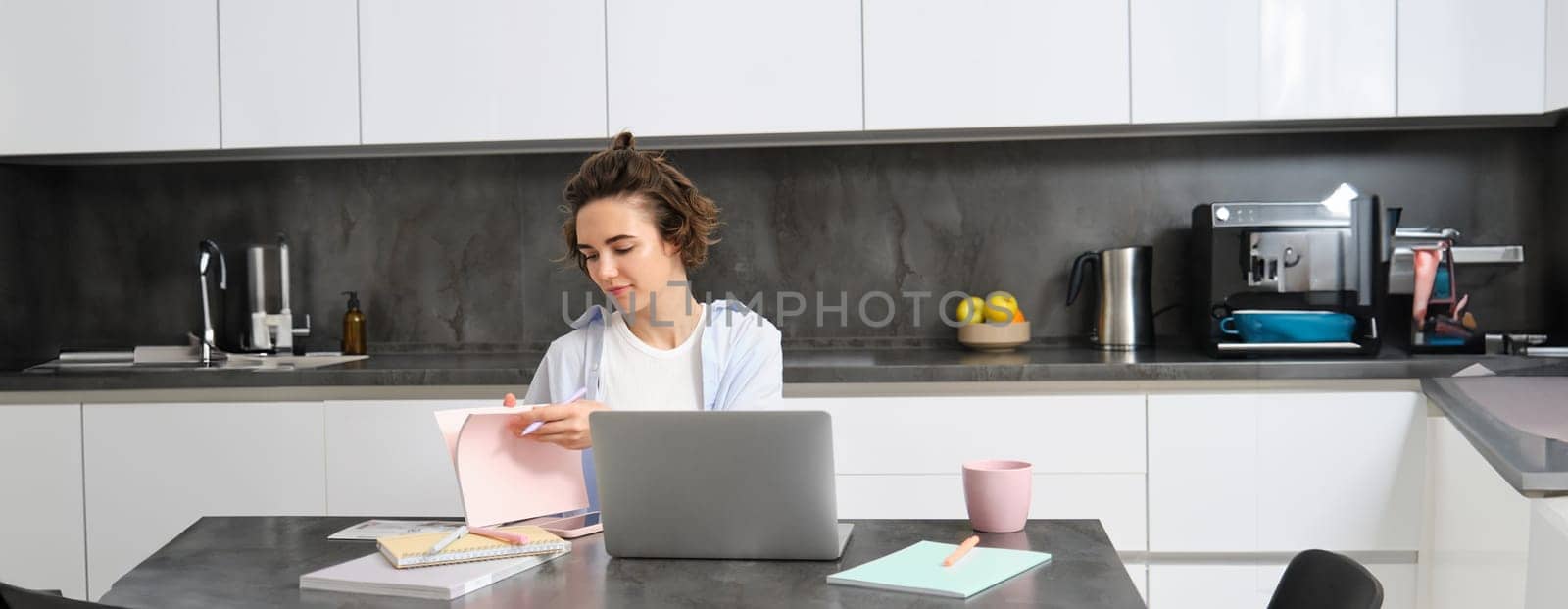 Portrait of working woman at home, checks her notebook, flips page, connects to online meeting via laptop. Girl learns programming remotely, studies course in internet.