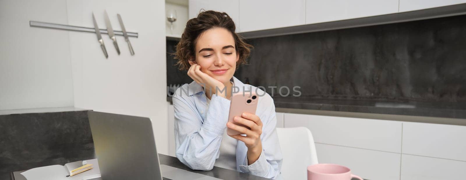 Image of young woman using her smartphone at home. Girl sits with mobile phone in kitchen and smiles.