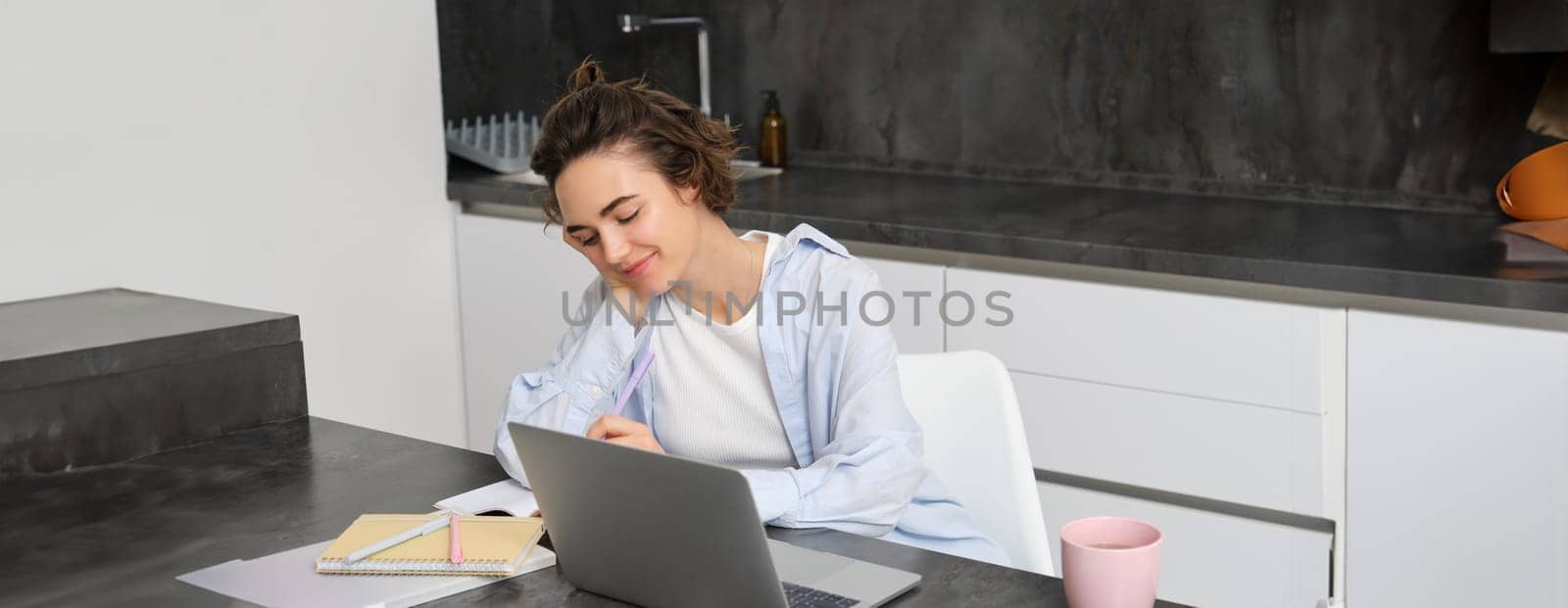 Image of young productive woman works from home, does distance learning, learns online course on her laptop in kitchen, writes down information, watches webinar, attends work meeting remotely by Benzoix