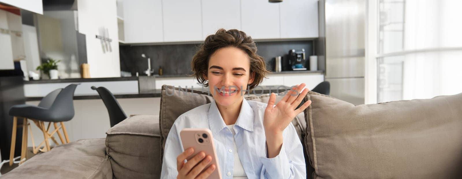 Lifestyle shot of happy girl talks on mobile phone, waves hand at smartphone camera, connects to online group chat on telephone app, sits on sofa at home.