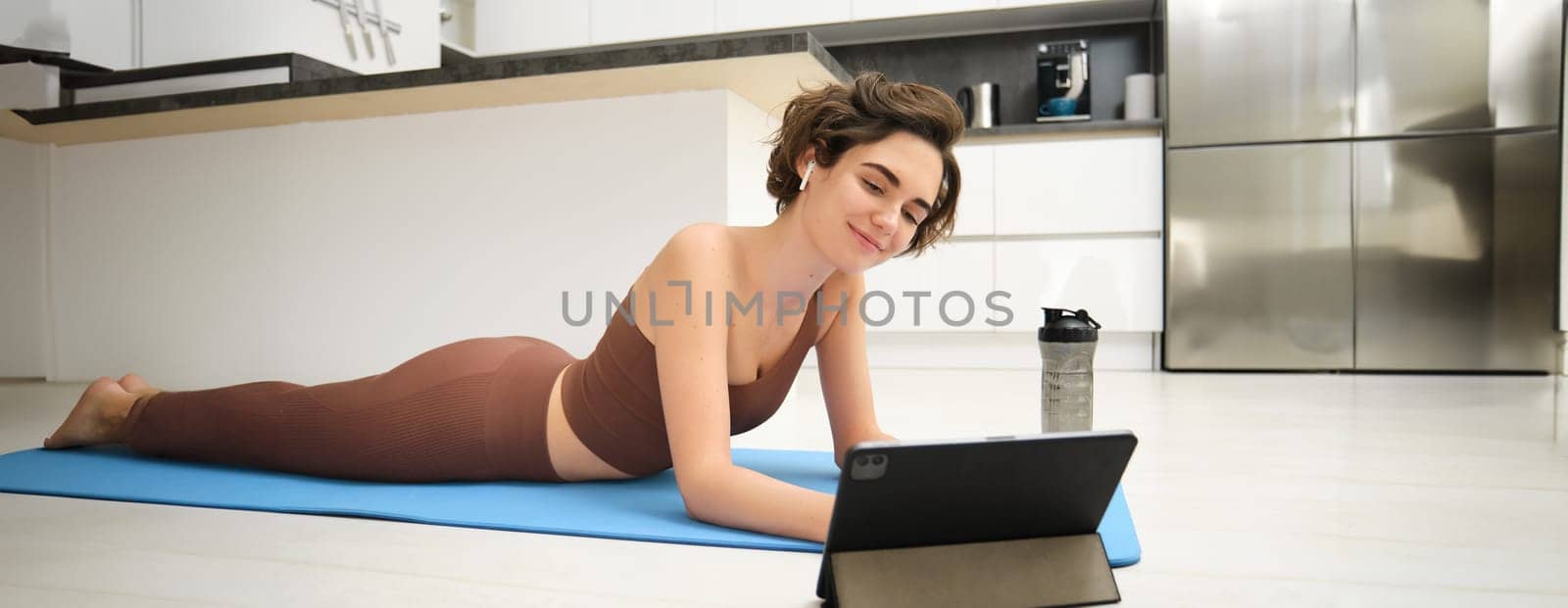 Full body smiling young strong sporty athletic fitness trainer instructor woman wear brown tracksuit use tablet for online training do exercises at home gym indoor. Workout sport concept by Benzoix