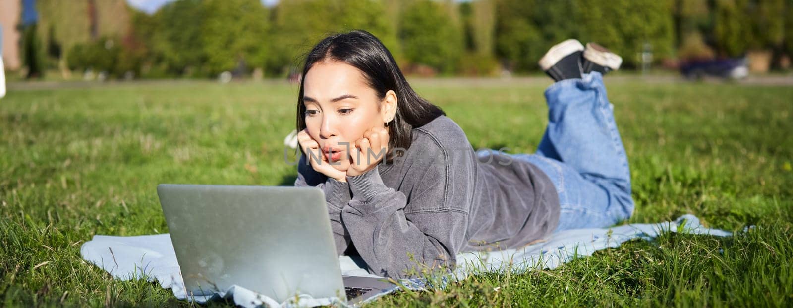 Portrait of beautiful girl lying in park and looking with surprised face at her laptop, watching videos, chatting with friends while relaxing outdoors.