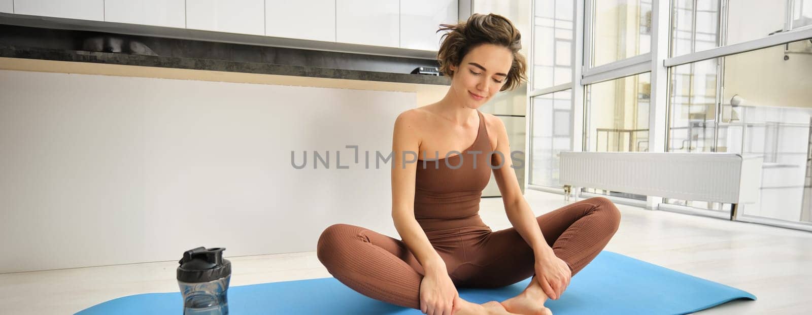 Sport and wellbeing. Young brunette fitness girl, wearing activewear, sitting on yoga mat, meditating, practice relaxation and mindfulness at home, workout in kitchen. Copy space