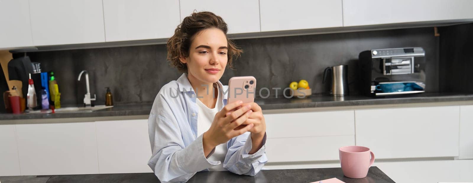 Portrait of beautiful smiling woman sitting at home with smartphone, using mobile phone app, orders groceries delivery.