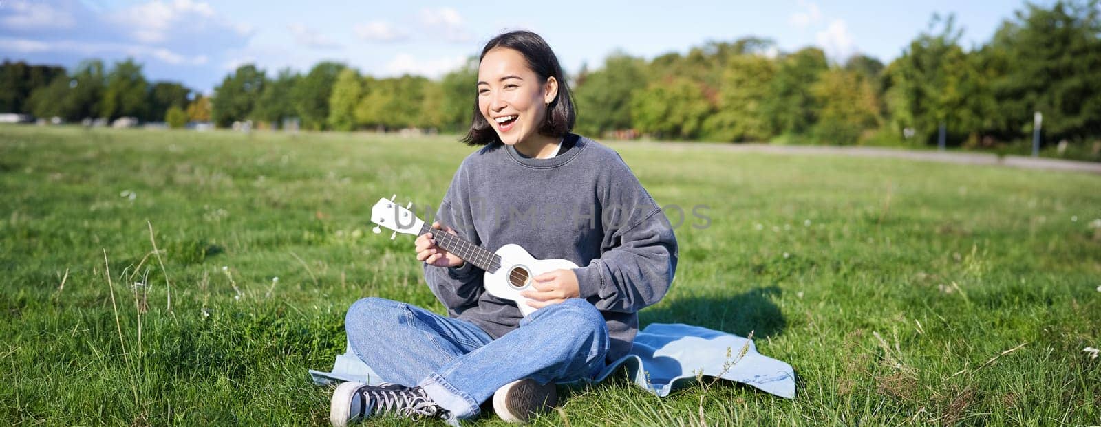 Happy brunette asian girl, student playing ukulele sitting on grass in park, relaxing, singing song, lifestyle concept.