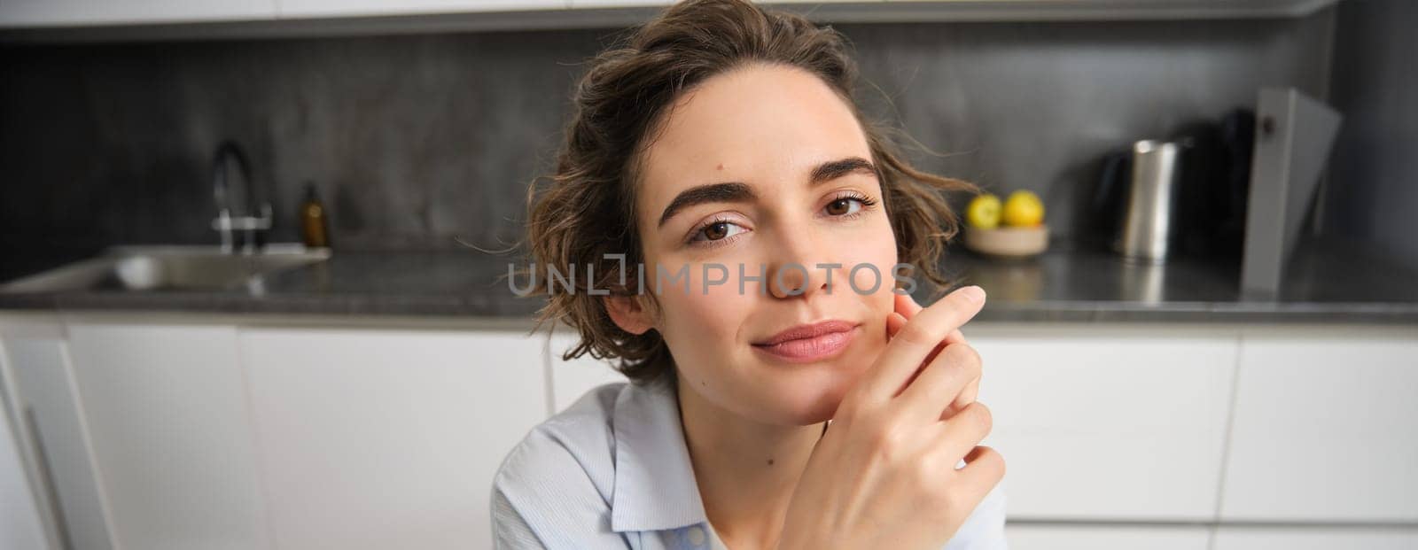 Close up portrait of young businesswoman, female entrepreneur, looking at camera and smiling with confidence, sitting at home in kitchen.