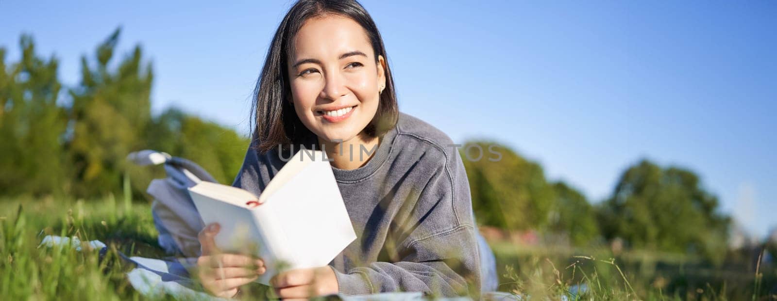 Portrait of cute korean girl, reading in park while lying on grass, relaxing with favorite book in hands, smiling happily.