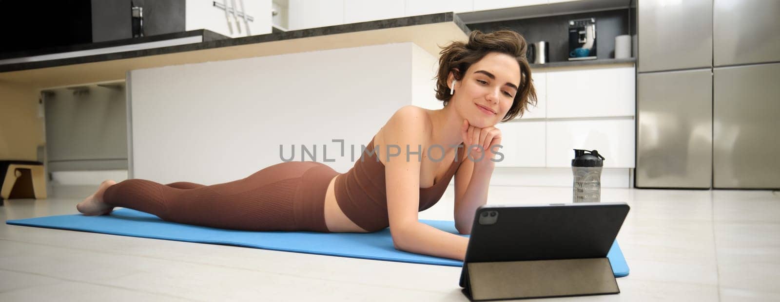 Portrait of smiling girl in fitness tracksuit, lying on mat at home, looks at tablet, watches online sports video tutorial, meditaiton lesson, yoga or workout online gym instructor by Benzoix