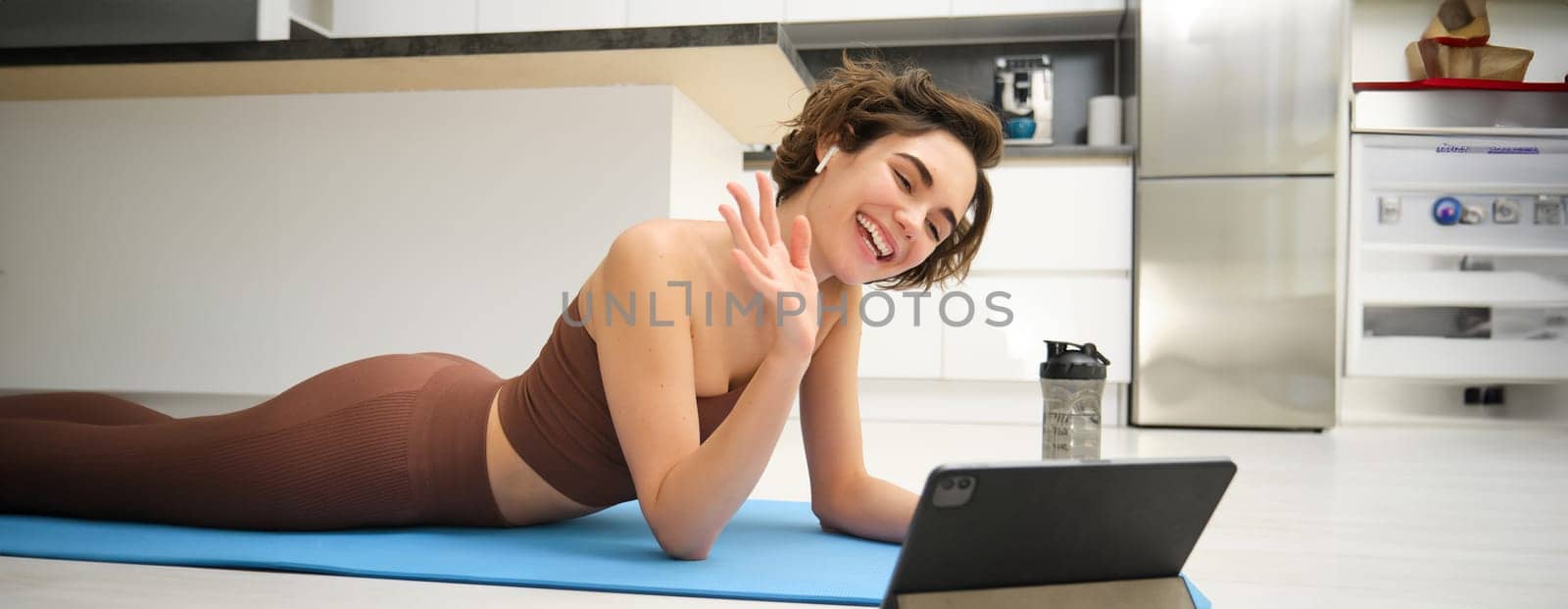 Sport and lifestyle. Young fitness woman, gym instructor waves hand at tablet, joins online video class, yoga and mindfulness workout, training from home indoors.