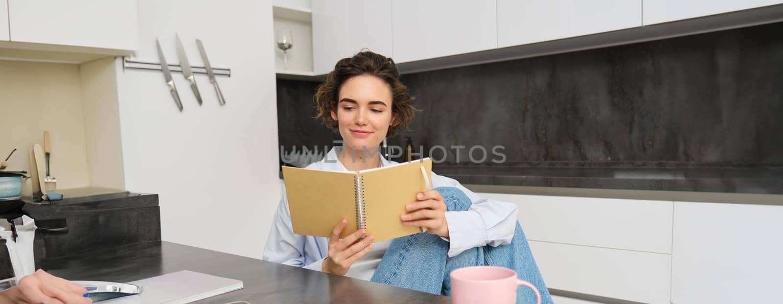 Young woman sitting in kitchen, reading her notes in notebook. Smiling girl at home, doing homework, studying by Benzoix