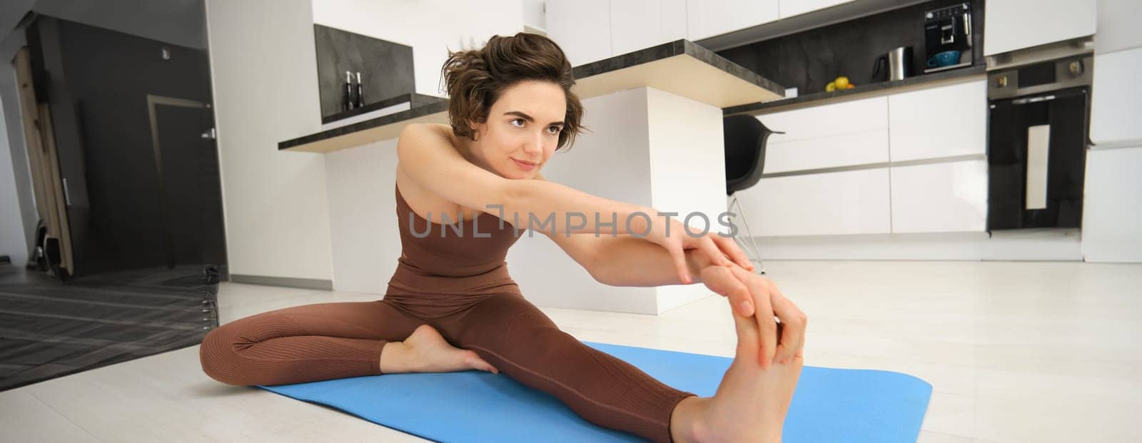 Portrait of young healthy woman stretches her legs, leaning towards feet, warm-up, doing workout exercises at home in bright room. Sport and lifestyle concept