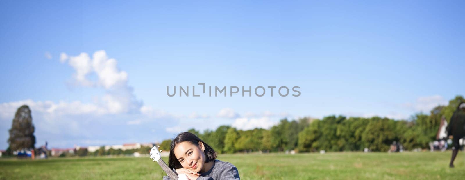 Romantic asian girl sitting with ukulele guitar in park and smiling, relaxing after university, enjoying day off on fresh air.