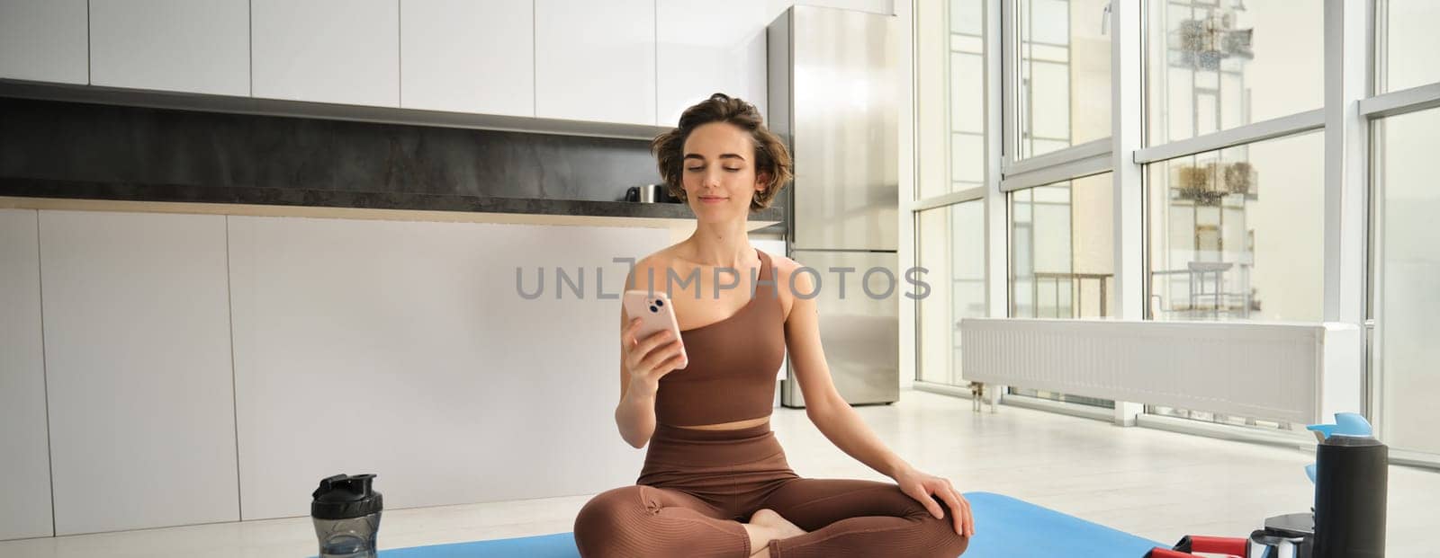 Sport app and wellbeing. Young woman, athlete sitting on floor at home with smartphone, doing yoga, using workout app on mobile phone, doing exercises at home. Copy space