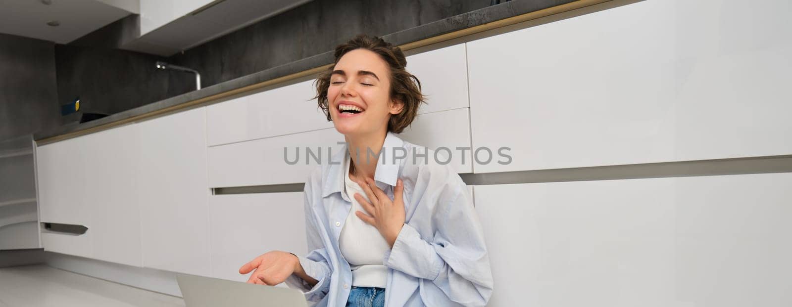 Portrait of young freelance girl connects to online meeting, video chats on laptop, sits on kitchen floor and enjoys her conversation.