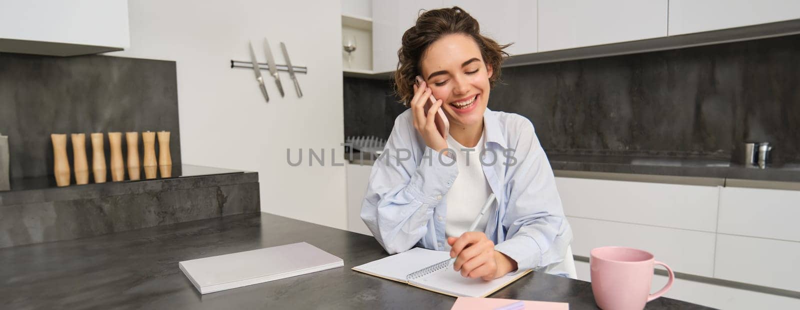 Image of woman working from home, writing down information during a phone call, calling someone, using smartphone.