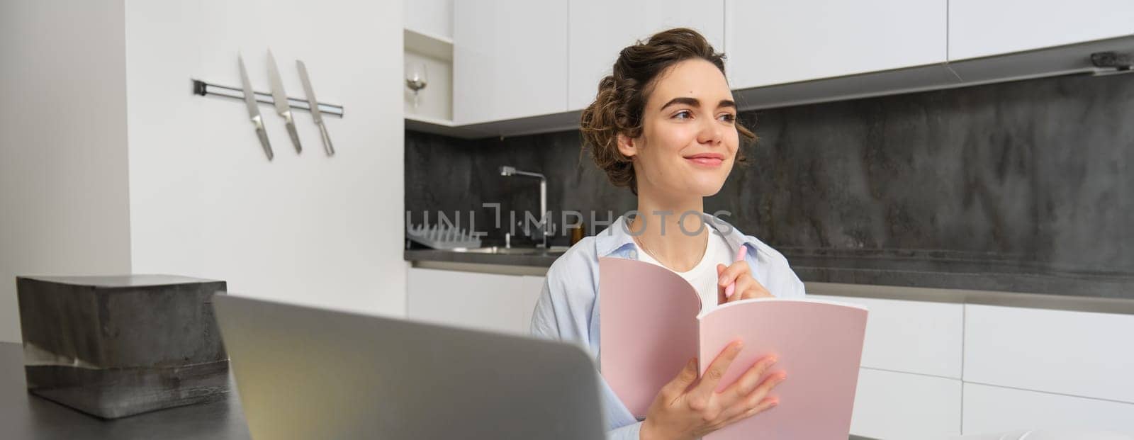 Young woman checking her schedule while working on remote from home, using laptop, looking at her daily planner, making notes, writing down information in notebook.